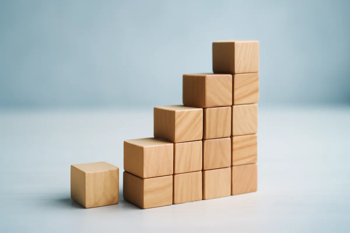 Wooden blocks stacked into a rising staircase against a pale background, with one loose block sitting apart from the stack
